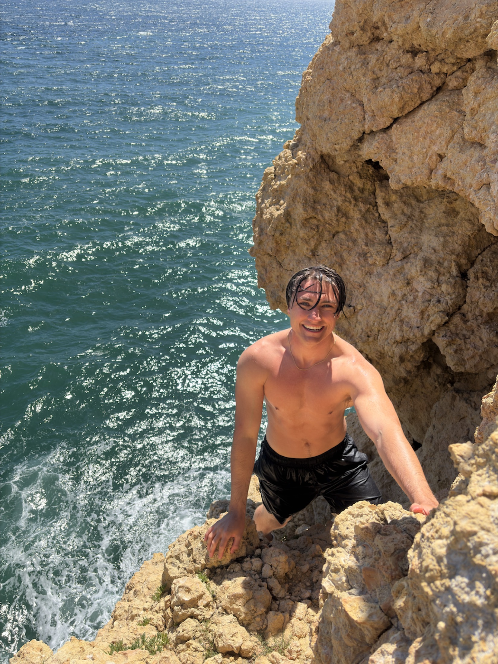 Angelo climbing back up a sunlit sea cliff above bright blue water
